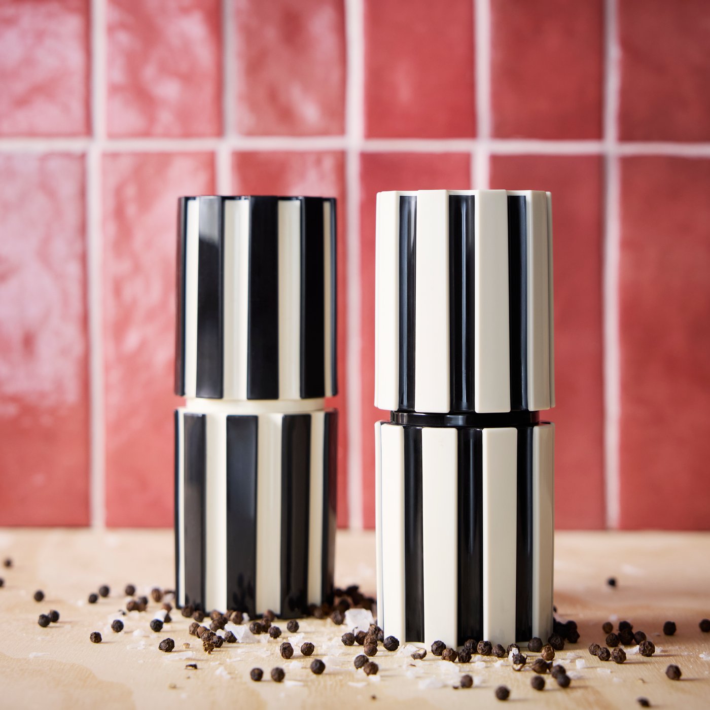Dviejų juodų / šviesiai smėlio spalvos SPJUTLILJA prieskonių malūnėlių rinkinys stovi ant paviršiaus, ant kurio išbarstyti juodieji pipirai ir druska.