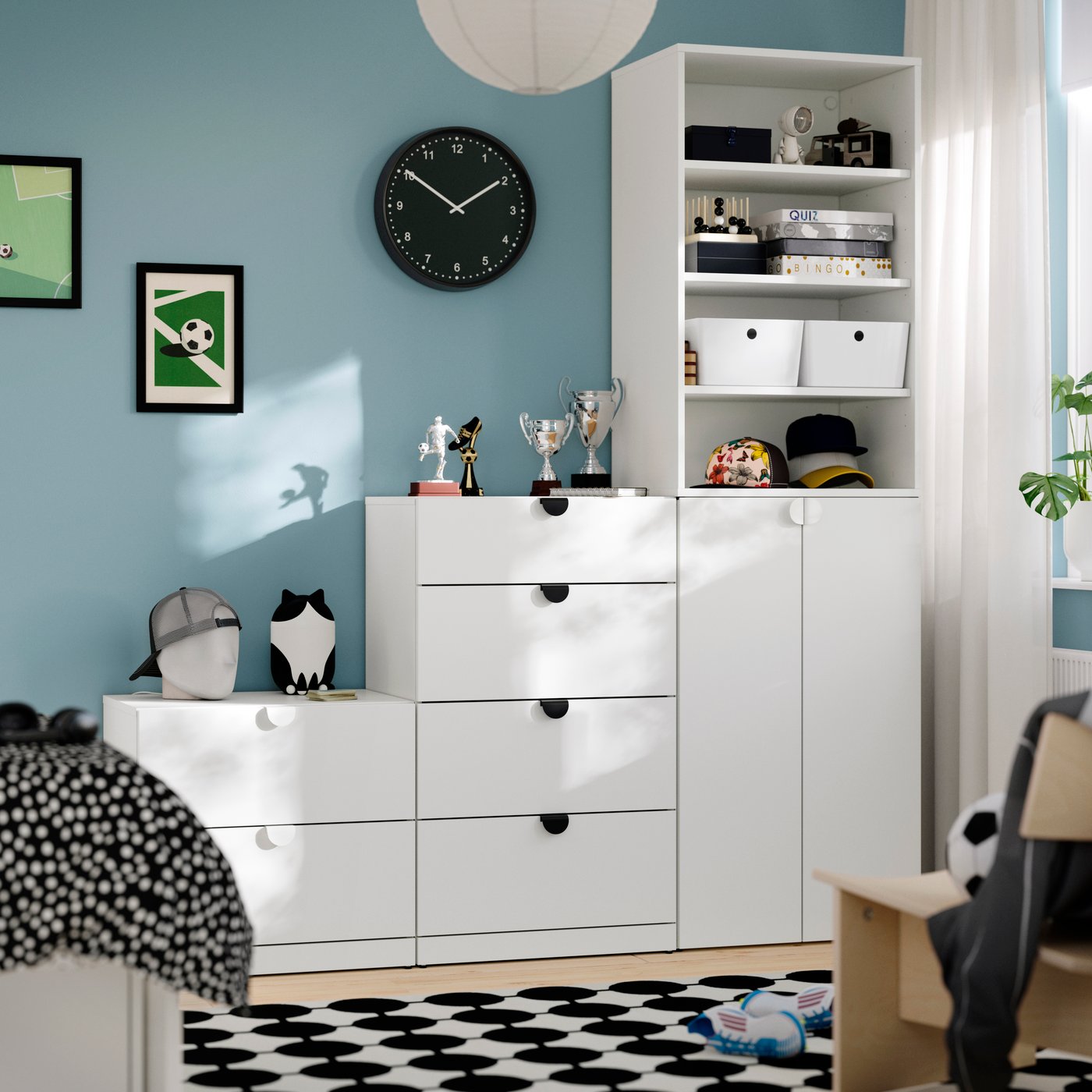 Bright child’s bedroom with a blue wall, white storage cabinets, a black wall clock, soccer-themed artwork and toys, sports trophies, and a patterned black-and-white rug. Sunlight streams through a nearby window.