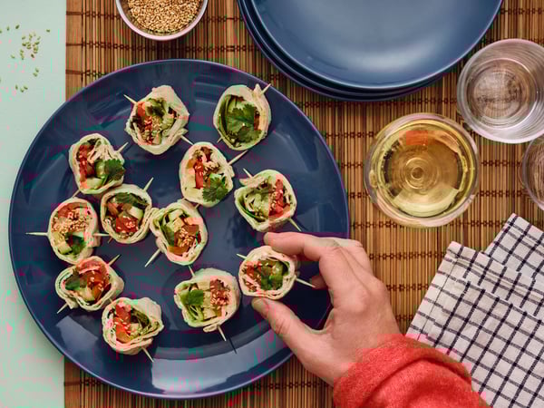 A hand reaches for a GRÄDDAT soft thin flatbread pinwheel with grilled peppers from a table with glasses and blue plates.