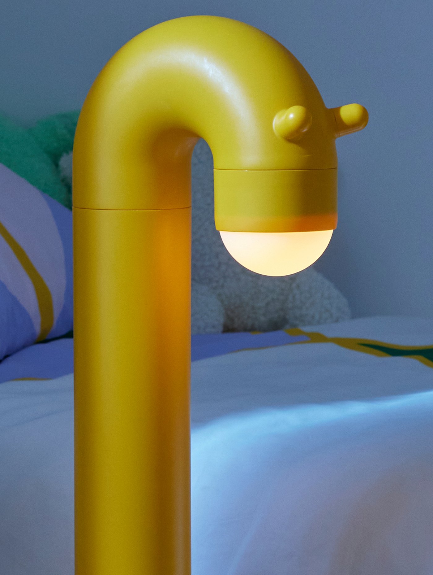 Żółta, zakrzywiona lampka nocna przypominająca postać przy łóżku.