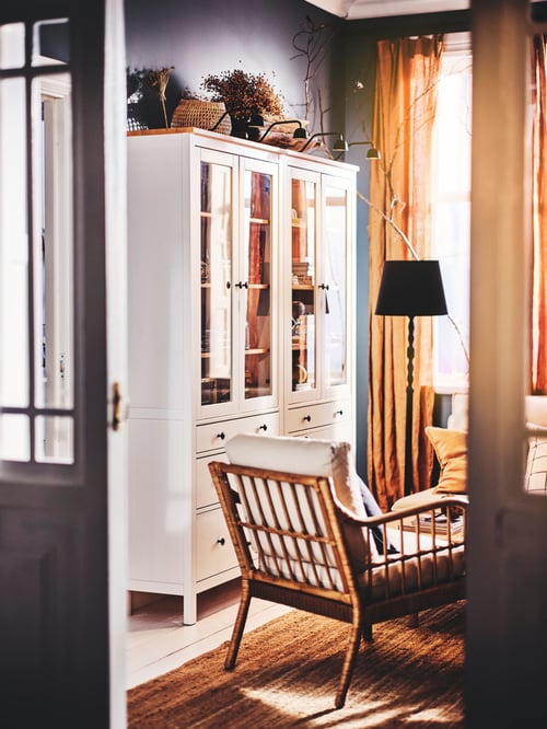 Living room with large windows, armchair, and tall white wooden glass door cabinet with 3 drawers holding decorations.