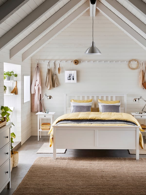 Ein weißes Schlafzimmer mit einem HEMNES Bett, Nachtkästchen und Kommode, sowie gelb / grauer Bettwäsche und einen hellbraunen Teppich.