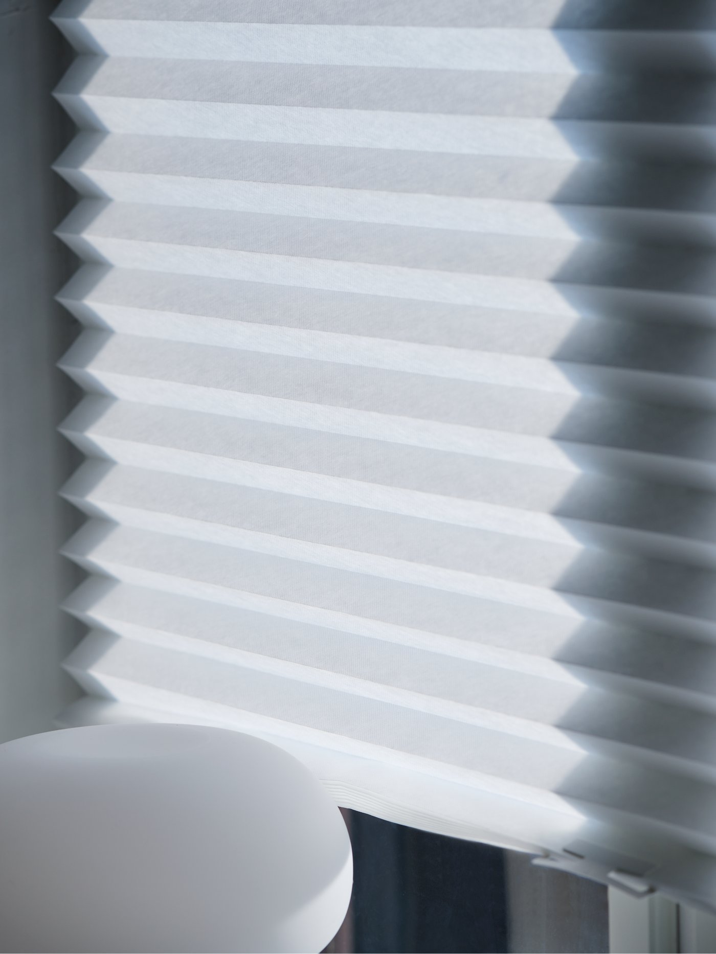 Detalle de un estor plisado blanco de poliéster junto a una lámpara de mesa beige y blanca.