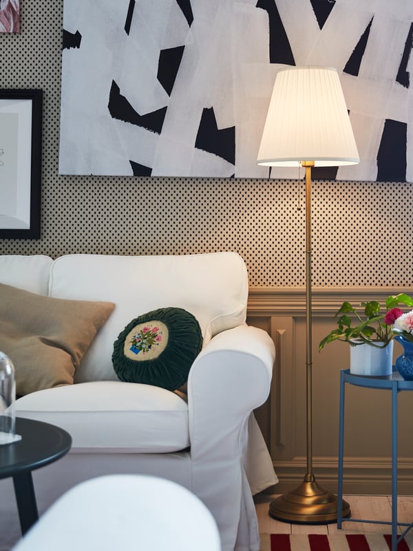A floor lamp in brass and white is lit and is standing next to a white EKTORP sofa in a living room.