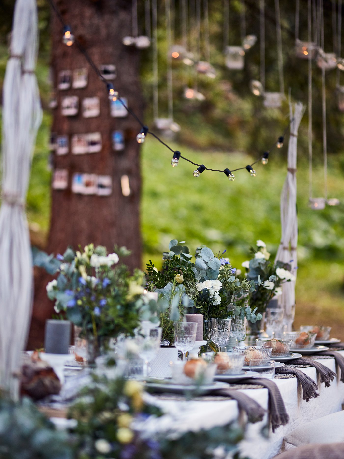 Et bord er pyntet op til fest udenfor, med blomster, lyskæder og service.
