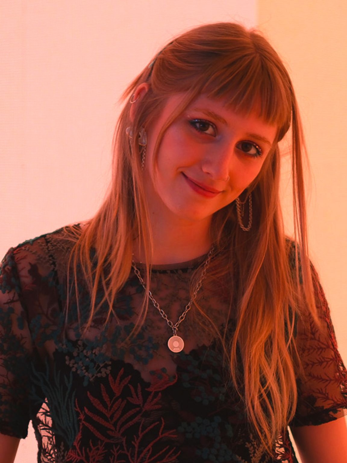 Rea, ki nosi črno majico in ogrlico, stoji na odru pred ozadjem nežno rdeče barve, za ozadje poskrbi digitalni zaslon, ki se med milanskim tednom oblikovanja 2024 uporablja na odru prizorišča IKEA. 