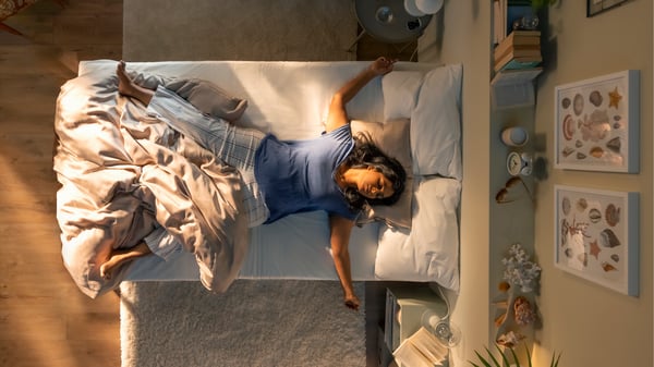 An aerial view of a person, sleeping on their back and sprawled across a bed in a warm, cosy-looking bedroom.