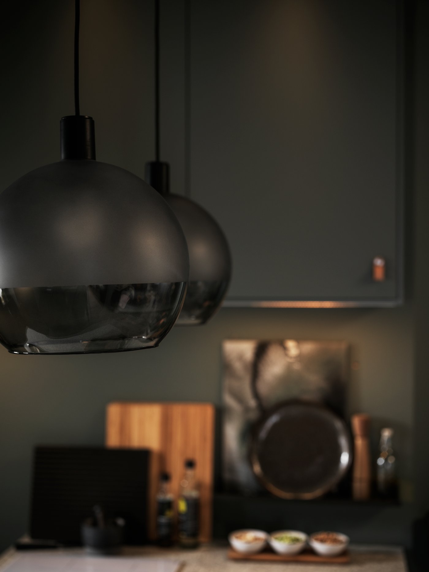 Två mörktonade pendellampor hänger ovanför en köksbänk med skärbrädor, kastruller och små skålar. Köket har mörka skåp och varm belysning.