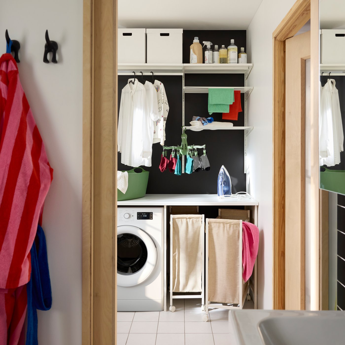 Vit ENHET tvättkombination med öppen förvaring, tvättmaskin, skjortor på galgar och två tvättpåsar på hjul.