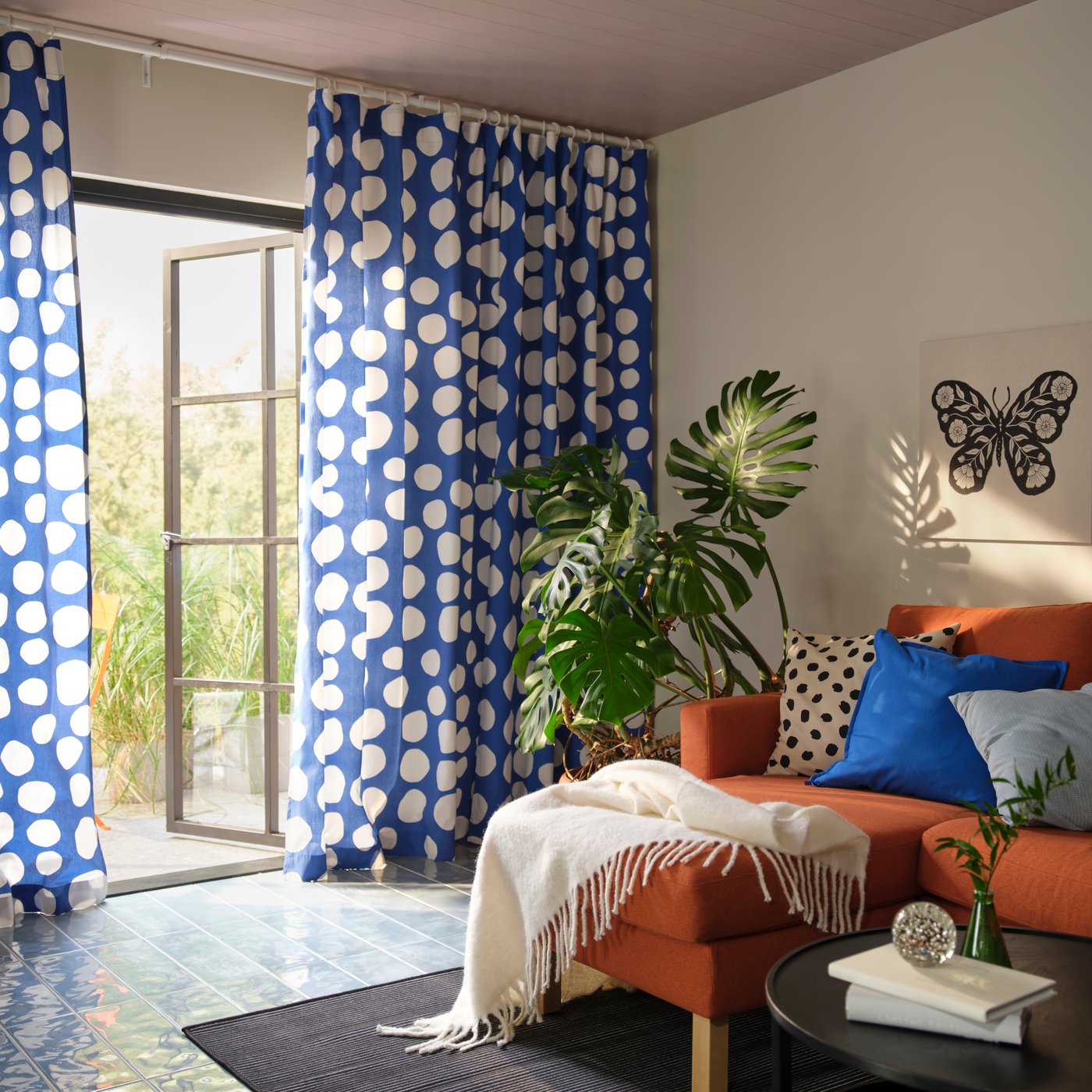 Ett vardagsrum med ett blåvitt RAKLOSTA förskuret tyg med runda vita prickar, tillsammans med gröna växter och en soffa.