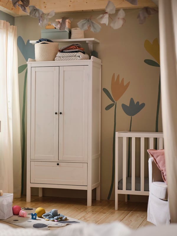 A white SUNDVIK wardrobe against a wall with a flower mural on it in a baby’s bedroom with a white SUNDVIK cot beside it.
