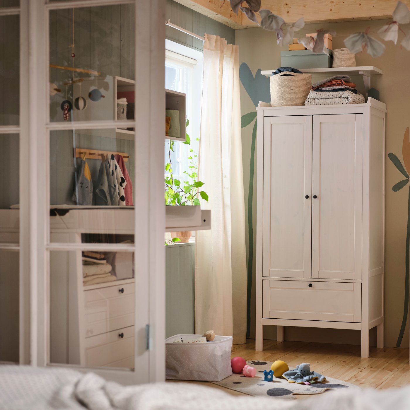 A white SUNDVIK wardrobe against a wall with a flower mural on it in a baby’s bedroom with a white SUNDVIK cot beside it.
