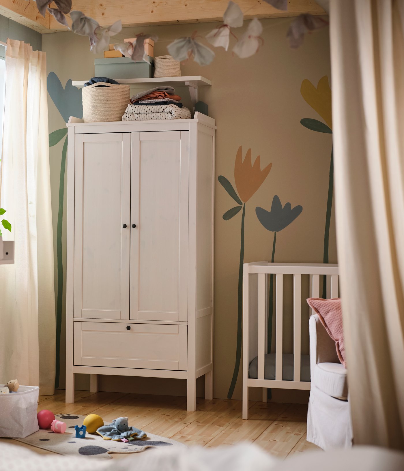 Un armadio bianco SUNDVIK addossato a una parete decorata con un murale floreale nella cameretta di un neonato, con accanto una culla bianca SUNDVIK.