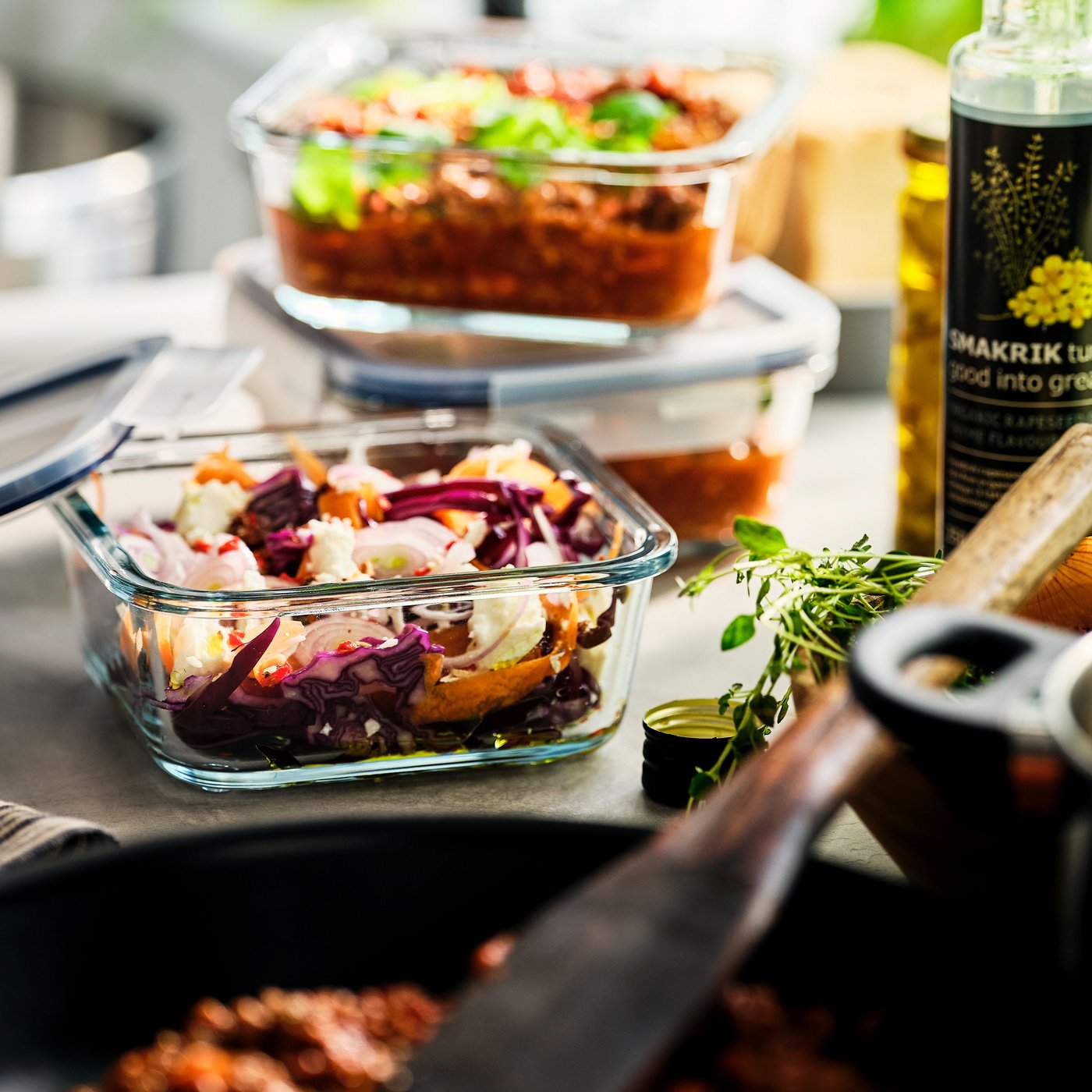 Een IKEA 365+ glazen voedselbewaarbakje met bereid voedsel staat naast een pan. Twee andere bakjes staan erachter gestapeld.