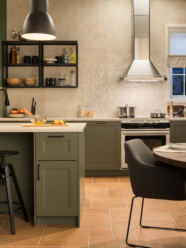 Stylish kitchen with AXSTAD green cabinets, open shelving, stainless hood, tiled backsplash, and dining area under warm pendant lighting.