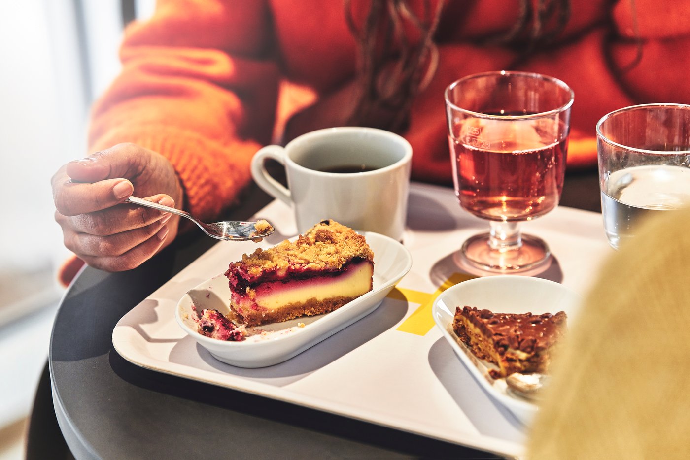 A person holds a spoon over a slice of berry cheesecake on a Swedish Restaurant table with coffee, soft drinks and another slice of cake nearby.