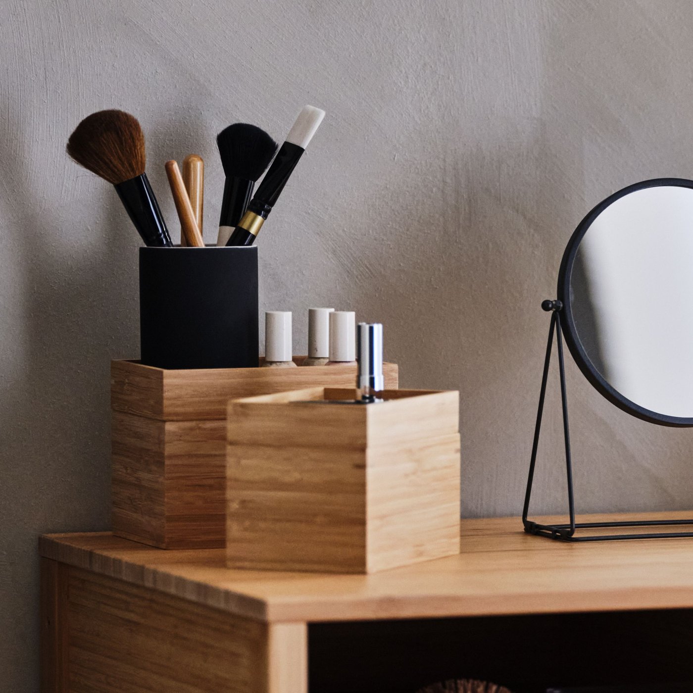 화장품과 메이크업 브러시가 담긴 대나무 소재의 보관용기 두 개가 테이블 거울 옆 목재 선반 위에 놓여 있는 모습.