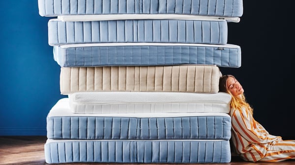 Une femme en pyjama rayé est appuyée contre une haute pile de neuf matelas de différentes nuances de bleu et textures, avec un matelas beige au milieu.