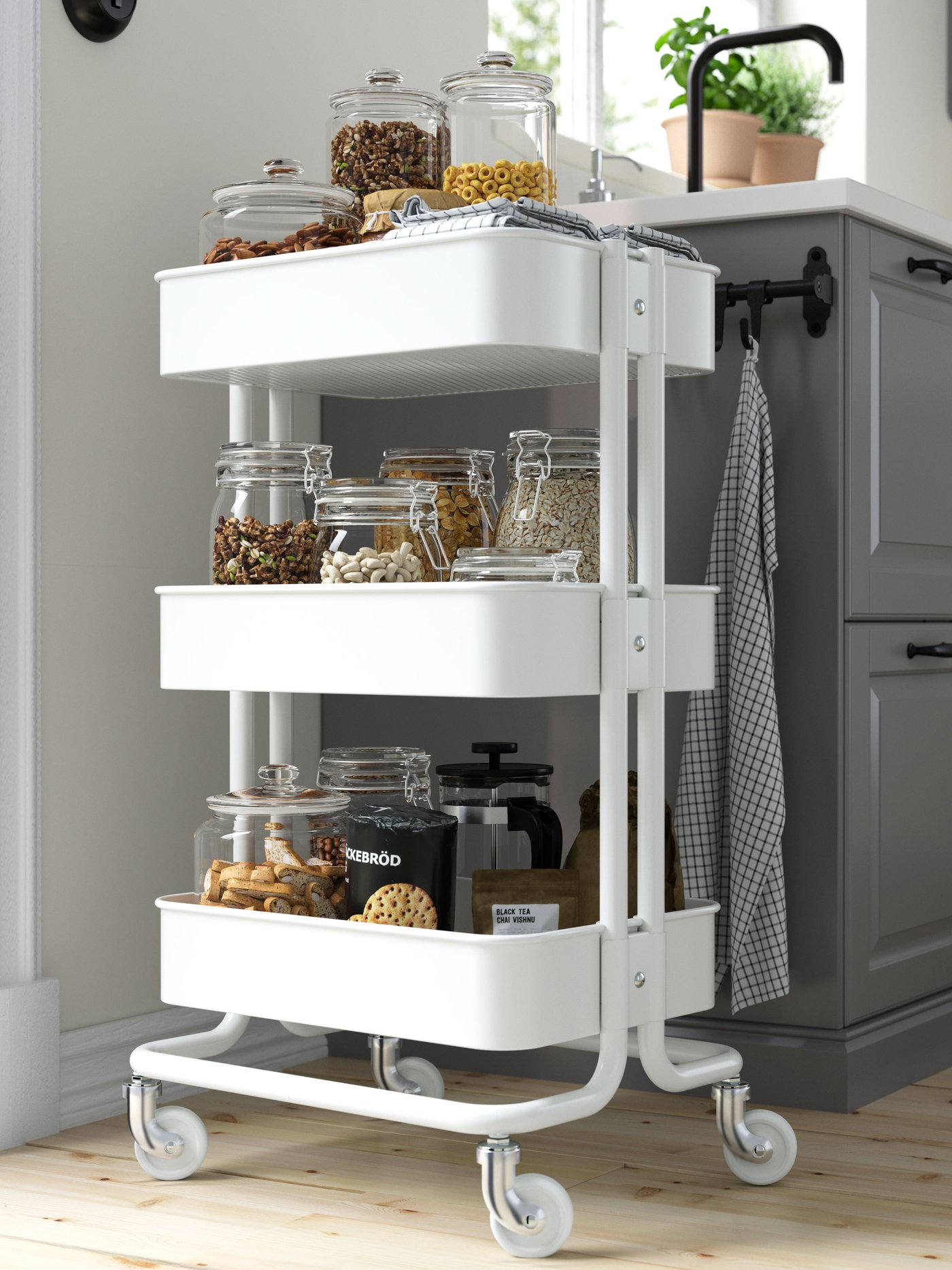 Un carrito de cocina RÅSKOG blanco al costado de un meson son frascos de vidrio en su interior.