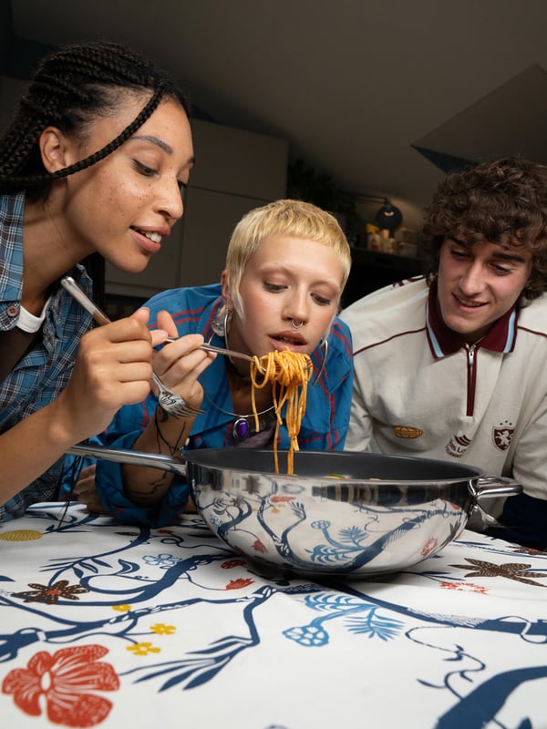 Tre giovani amici mangiano con allegria un piatto di spaghetti direttamente dalla pentola, condivisa al centro del tavolo.