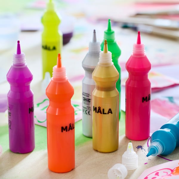 Close-up of 8 bottles of MÅLA fluorescent paint with lots of different paintings and drawings around them on the table.