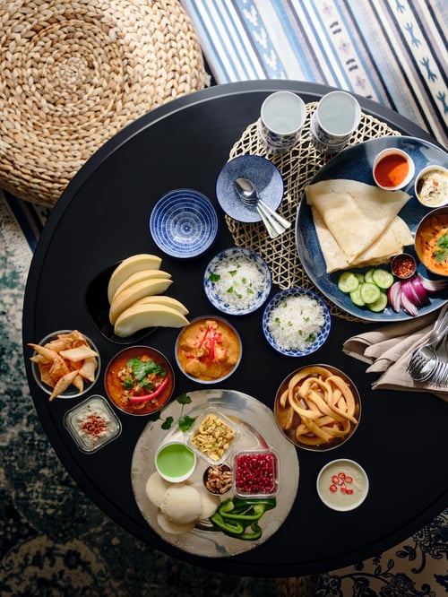 Vedere de sus a mesei rotunde pline de alimente, boluri de porțelan cu model albastru, cani albastre, scaun bej, covor albastru cu dungi mici.