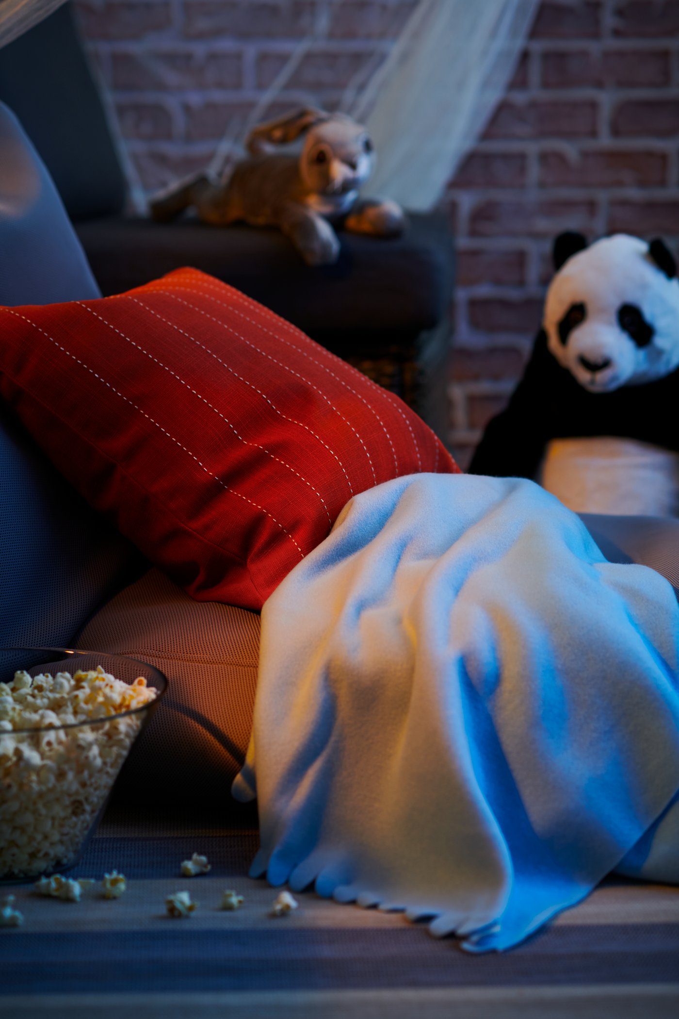 A hut made with cushions, throws, and cuddly toys. On the floor sits a glass bowl filled with popcorn.