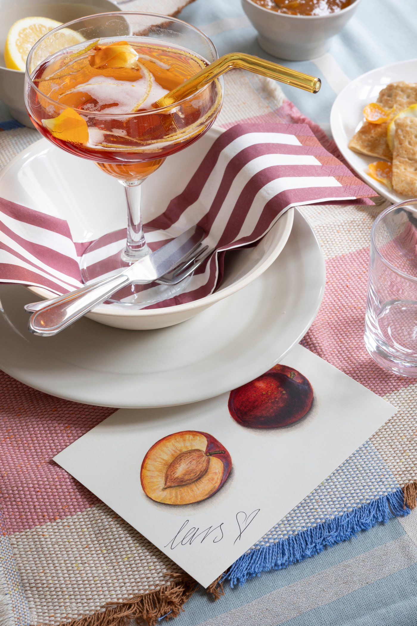 A white plate and large bowl dressed with a pink and white napkin, a champagne coupe on top, on a dining table with a pink, patterned place mat and a signed card placed on the side.