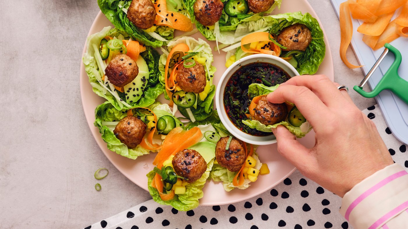 A hand is holding a meatball wrapped in a lettuce leaf over a pink plate and is dipping it into a white dish with soy sauce.