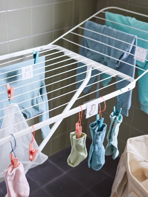 A white JÄLL drying rack with children’s clothes and socks hanging from SLIBB hanging clothes pegs with plastic loops.