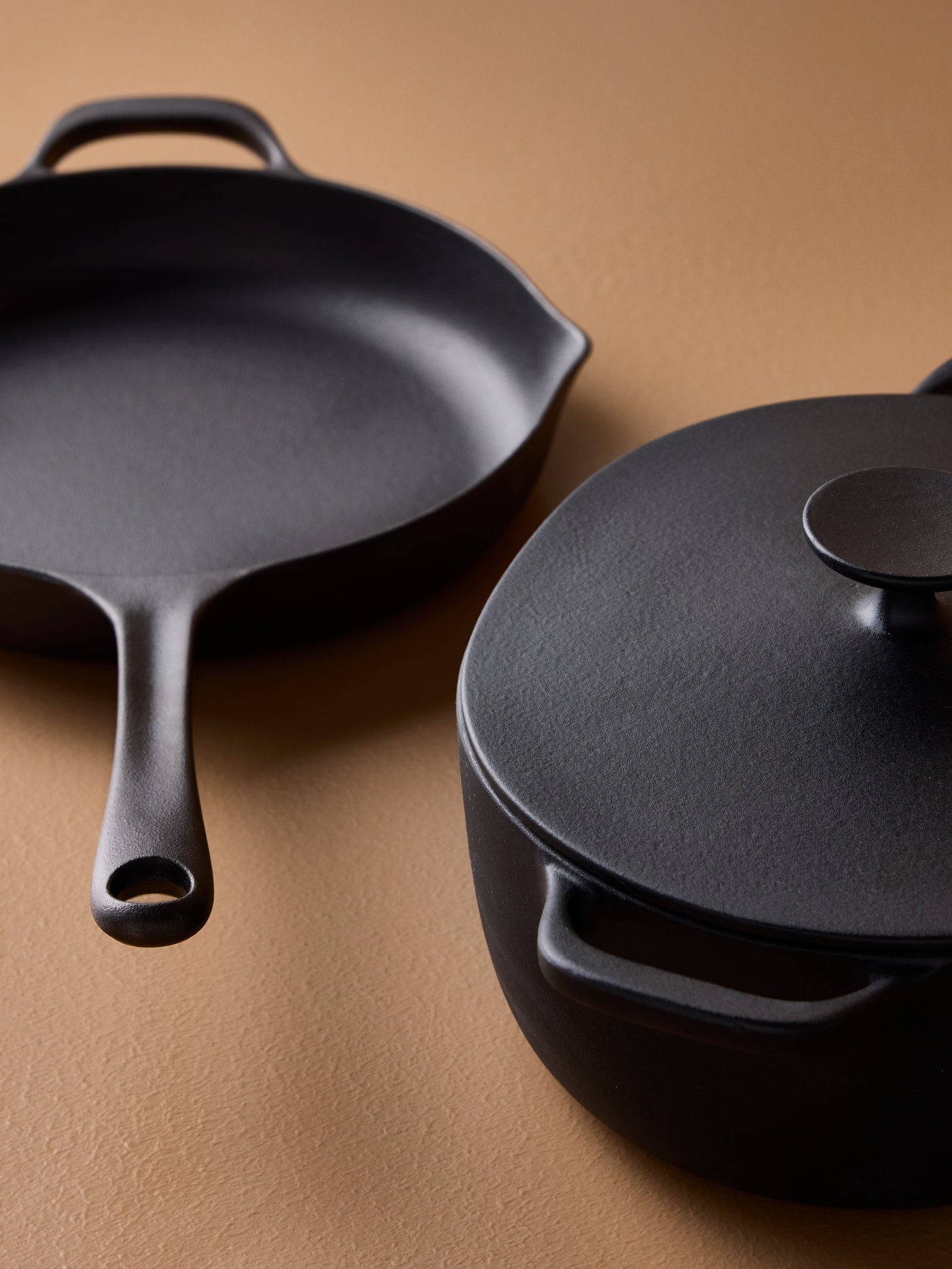 A VARDAGEN frying pan in cast iron and a VARDAGEN casserole with lid in enamelled cast iron are placed on a brown surface.