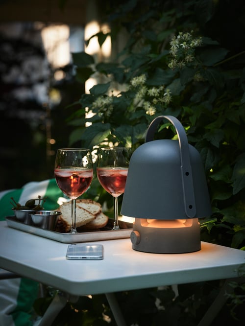Zonă în aer liber cu  o lampă cu boxă Bluetooth VAPPEBY aprinsă așezată pe o masă TORPARÖ lângă două pahare SVALKA cu lichid roșu.