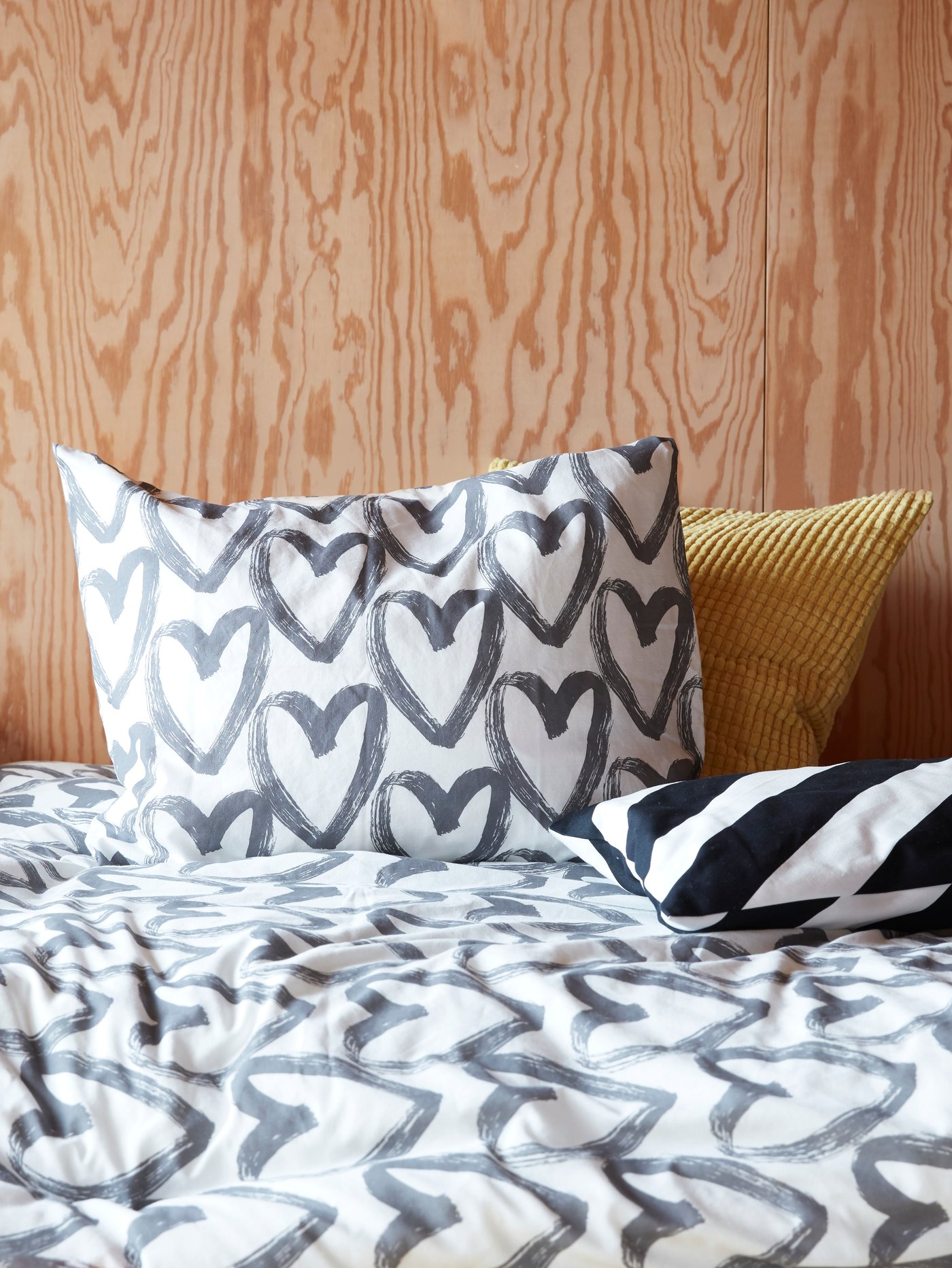 Ein Bett mit zwei Kissen und der LYKTFIBBLA Bettwäsche mit grauen Herzen auf einem weißen Hintergrund.