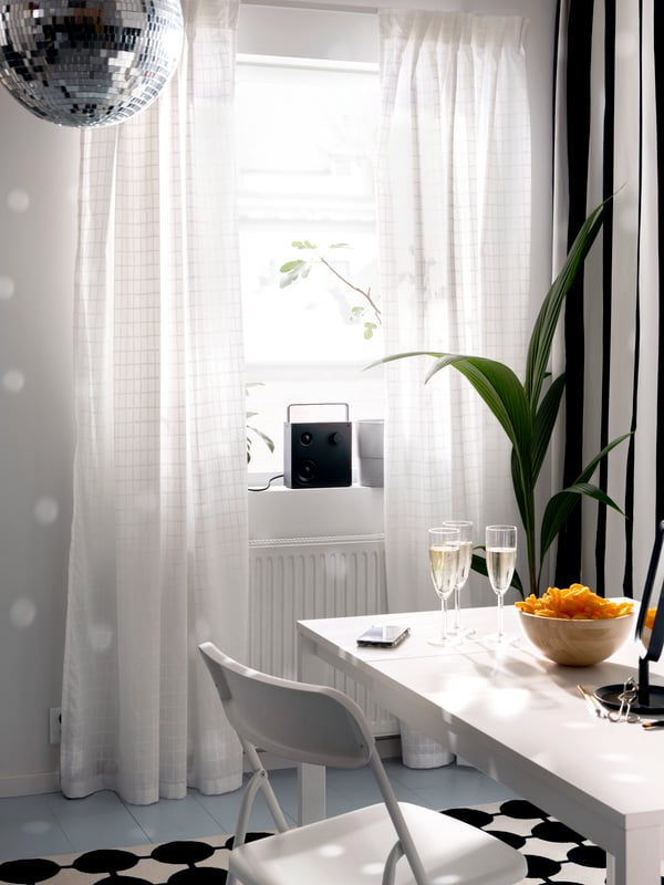 A VAPPEBY Bluetooth speaker stands in a window with MÄSTERROT curtains near a VIHALS extendable table and a folding chair.