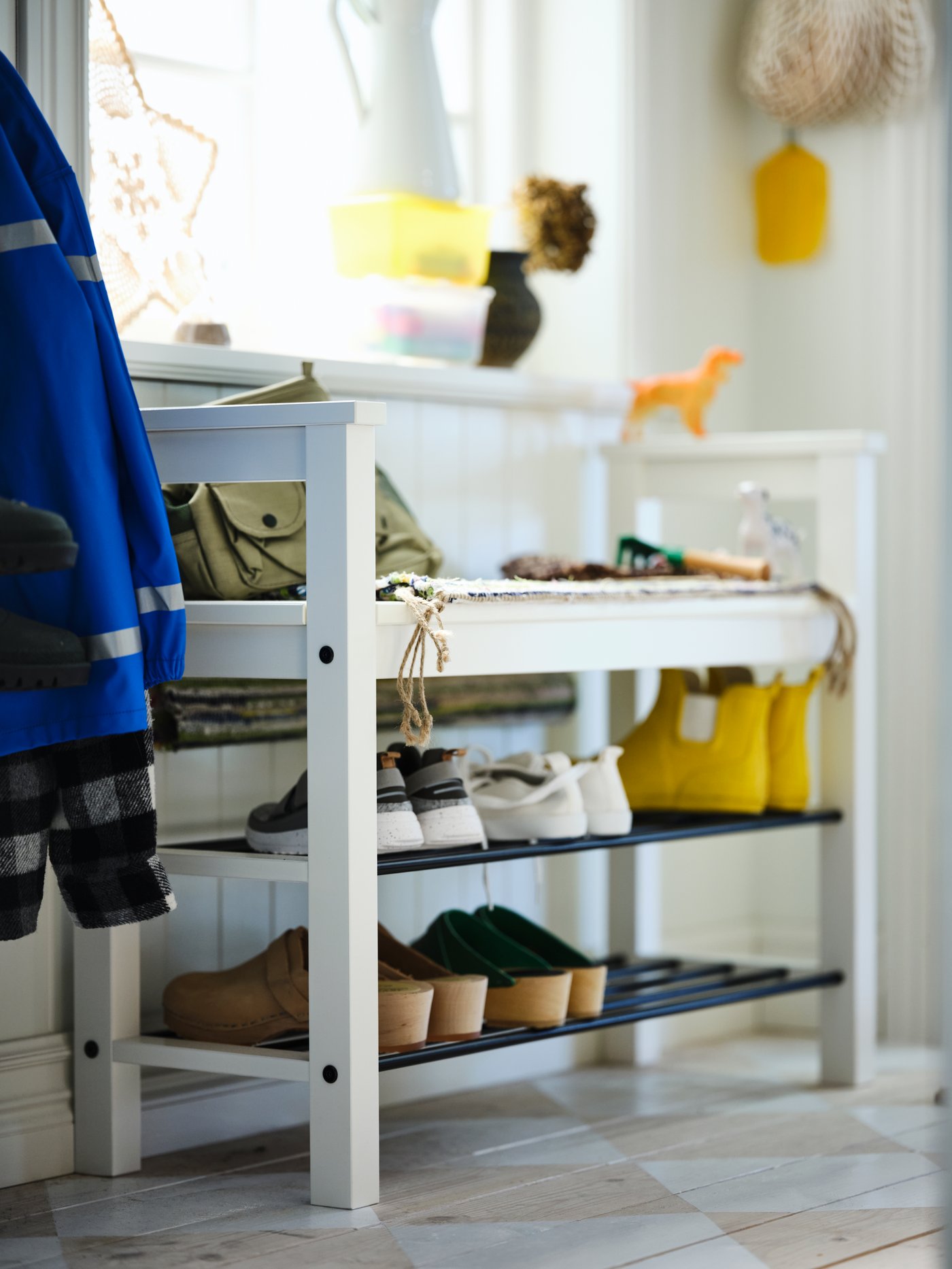 Ein weißes Schuhregal mit einer Sitzbank. Es stehen Schuhe im Regal, daneben hängen Jacken.