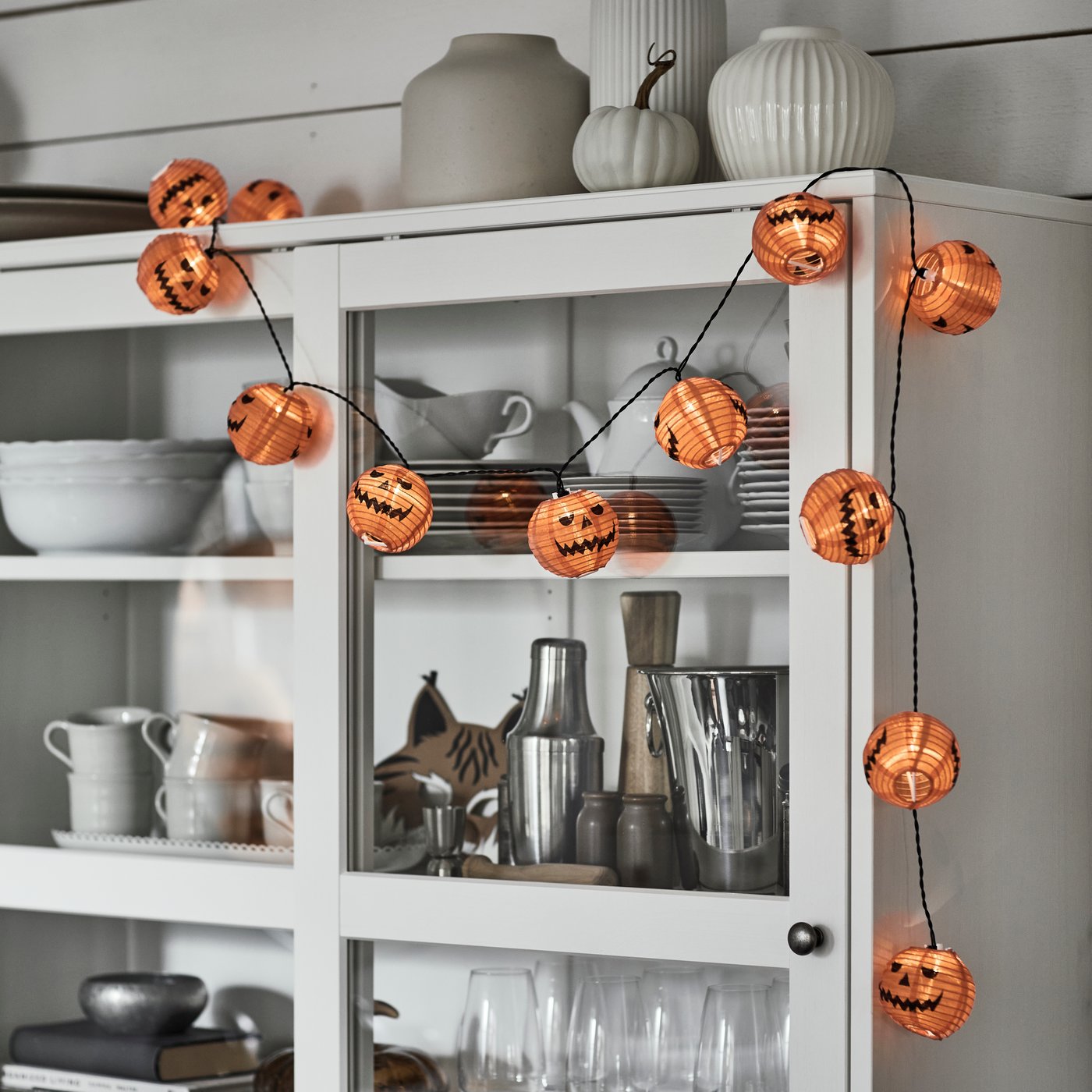 A KUSTFYR light chain featuring several small carved pumpkins is placed on top of a white cabinet with glass doors.