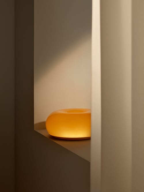 O veioză portocalie cu LED VARMBLIXT cu design rotund se află pe o suprafață gri închis. O sursă de lumină se aprinde din dreapta.