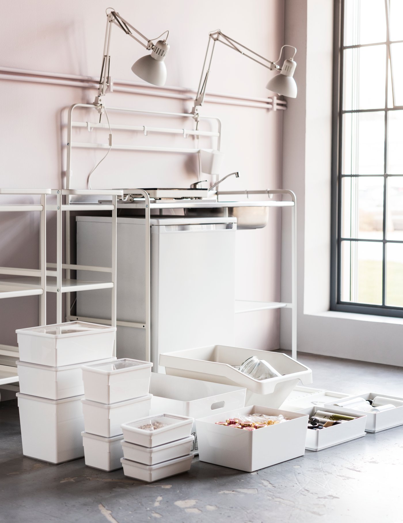 A bare white mini-kitchen with a fridge and two white work lamps. White waste sorting bins and boxes are on the floor.