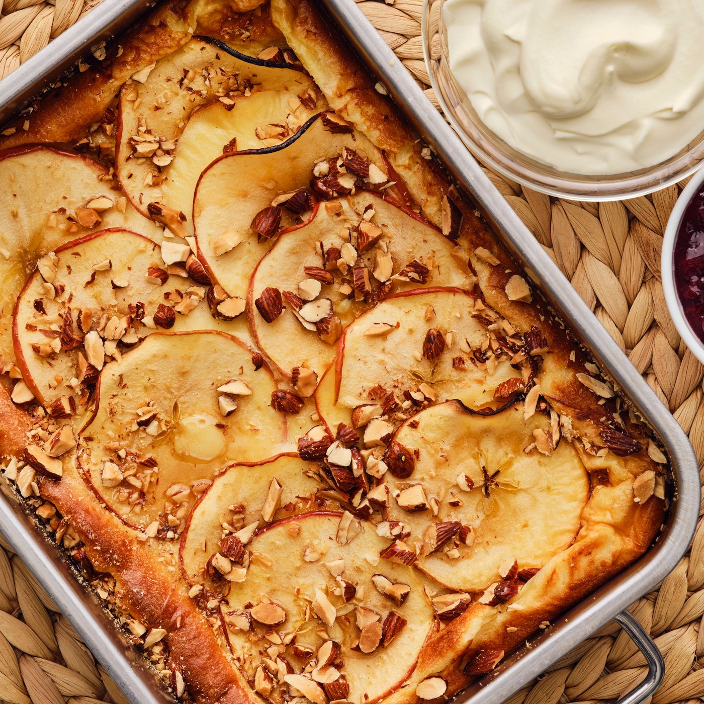 A golden-brown oven pancake with apples and almonds has been baked in a metal baking tray that is placed on a green surface.