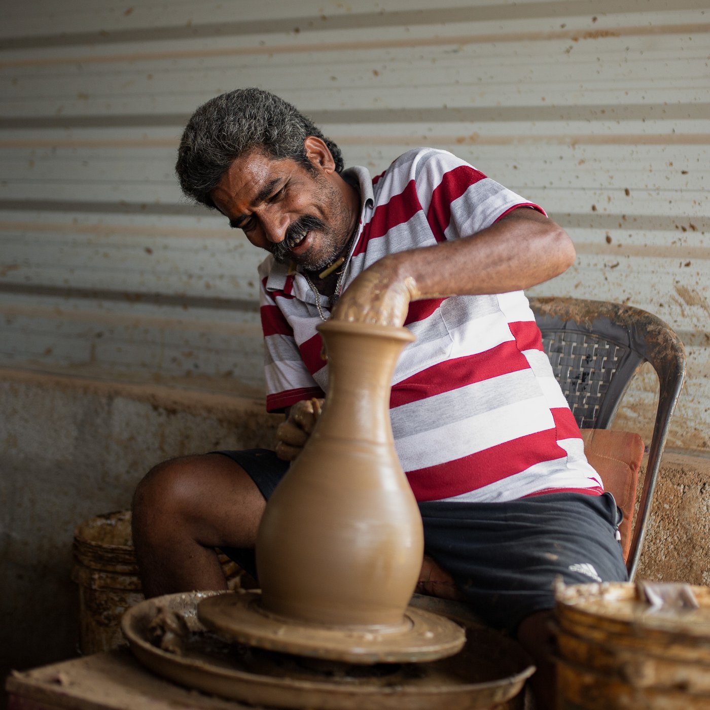Osoba radi za grnčarskim točkom i oblikuje visoku glinenu posudu. Osoba nosi crveno-belu prugastu majicu, sedi na stolici i koncentriše se na glinenu posudu, nadvijajući se iznad nje. U pozadini se vidi rustično okruženje s rebrastim metalnim zidovima, što ukazuje na to da je u pitanju zanatska radionica.