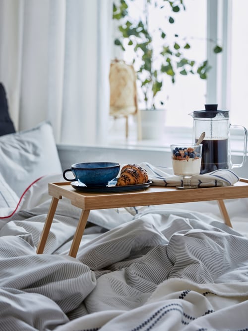 Bedroom with bed frame, bed tray, coffee maker, goblet, mug, saucer, bedding sets, table lamp, croissant, plant, curtains.