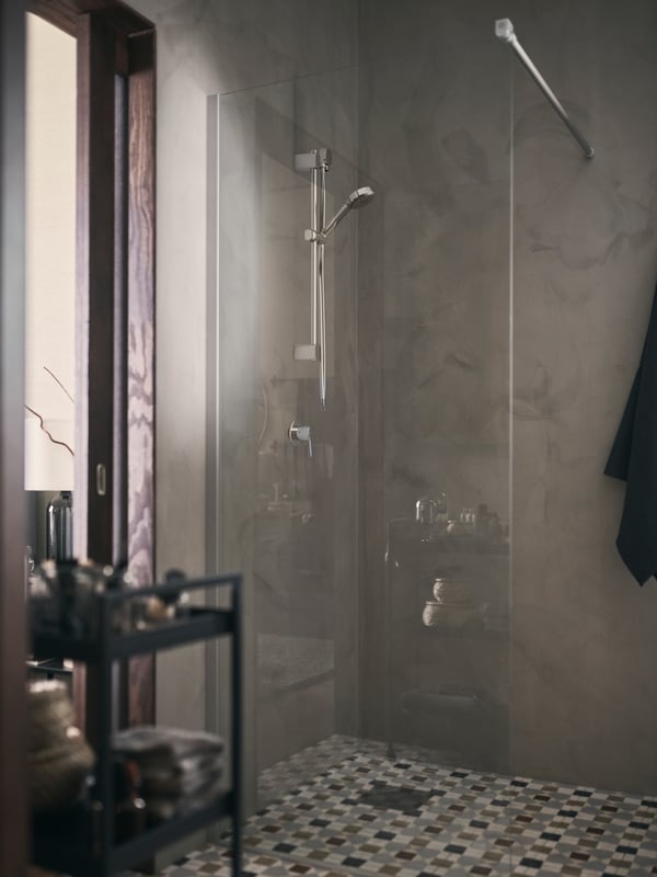 A walk-in BROGRUND shower with a glass screen beside a window, in the corner of a bathroom with a checked tile floor.