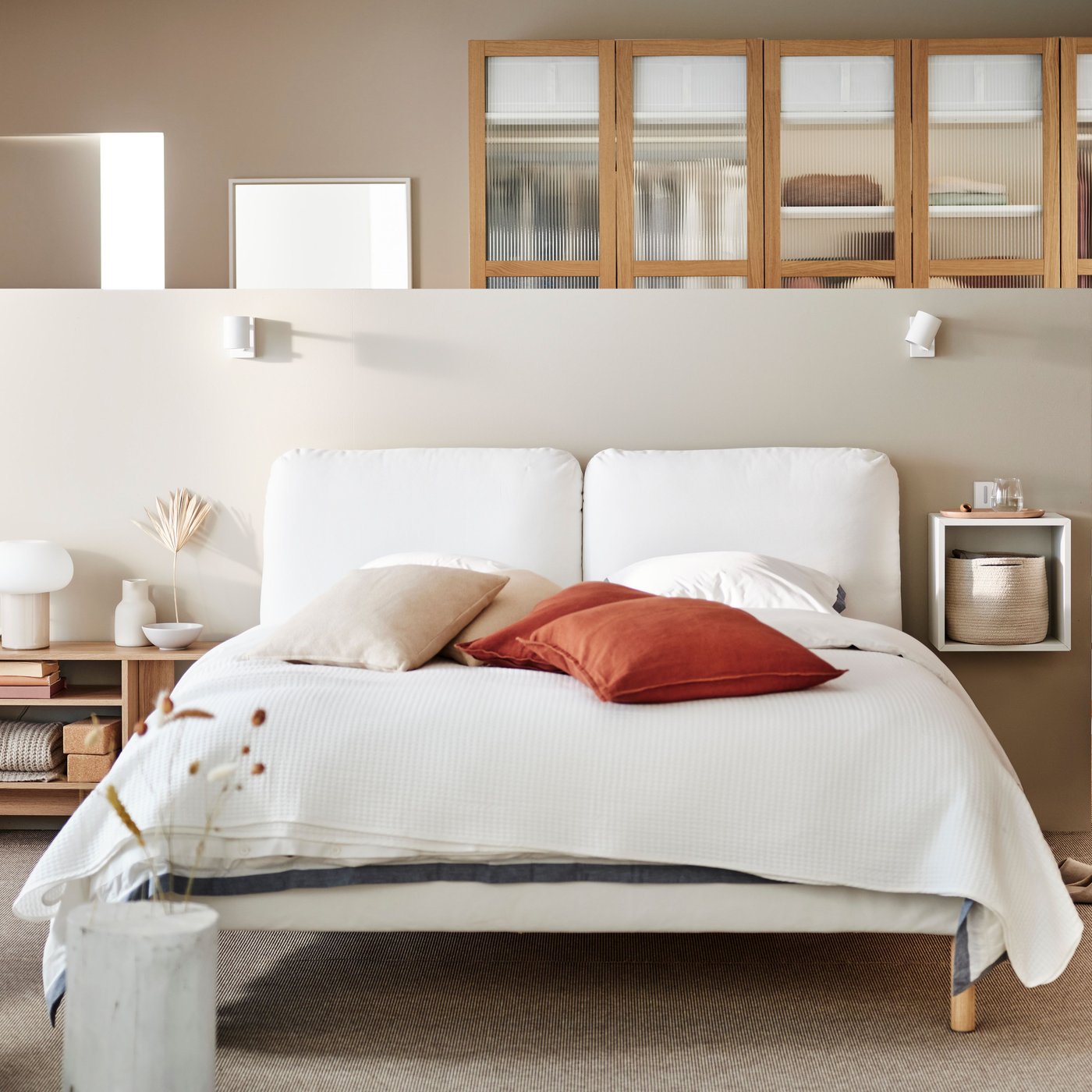 A SAGESUND upholstered bed in Diseröd brown/Luröy in a large airy bedroom covered in white bed linen with red cushions on it.