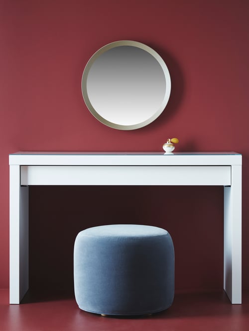 Bedroom with red walls, white dressing table, round mirror mounted on wall, grey pouffe, perfume bottle.