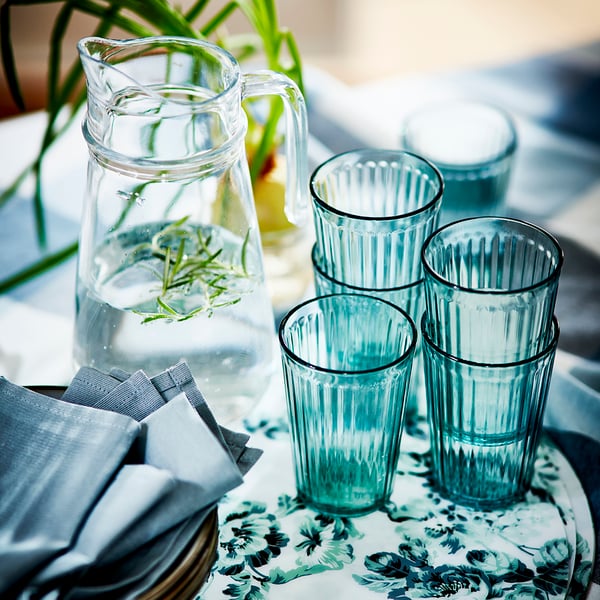 A green, vertically ribbed glass pitcher on a table with two clear glasses of water, one containing a lemon slice. The background features a blurred red brick wall, suggesting an indoor or patio setting. The scene conveys a refreshing atmosphere, possibly for a casual dining experience or home environment.