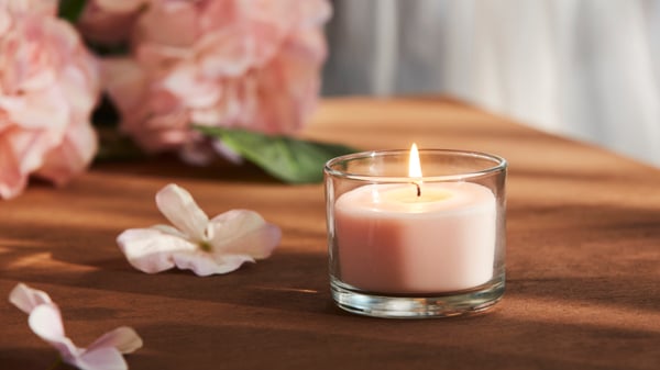 Eine rosa LOTSFAGEL Duftkerze ist auf einem Tisch zu sehen, im Hintergrund eine Künstliche SMYCKA Hortensie in Hellrosa, geeignet für den Innen‑ und Außenbereich