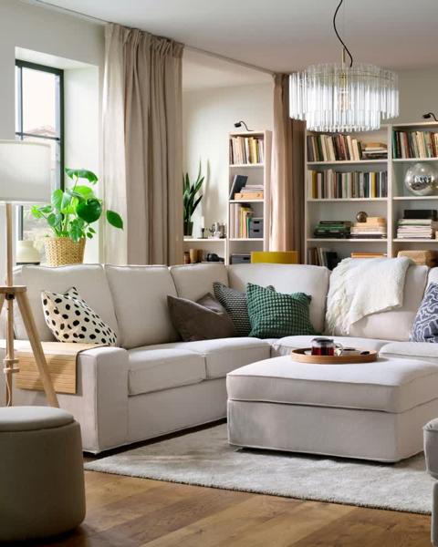 Ein helles Wohnzimmer mit einem hellbeigen KIVIK-Ecksofa, umgeben von Bücherregalen und dekorativen Pflanzen.