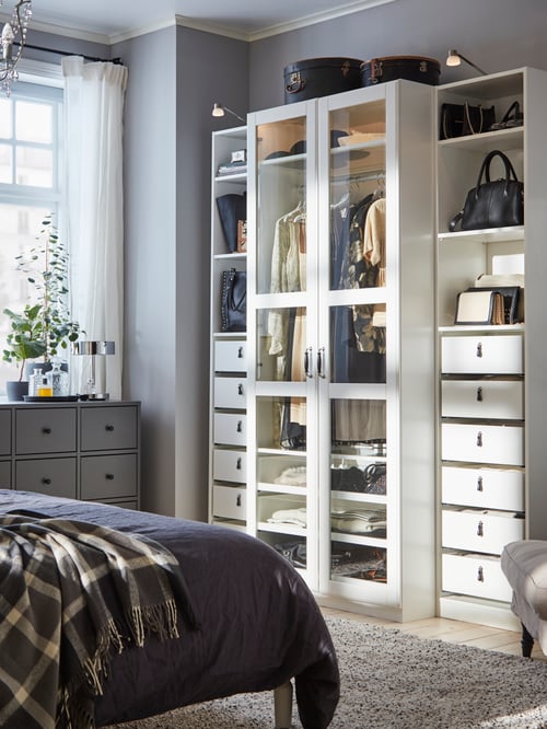 A bedroom has a tall white PAX wardrobe with glass doors in the centre. On each side are six white drawers and three shelves.