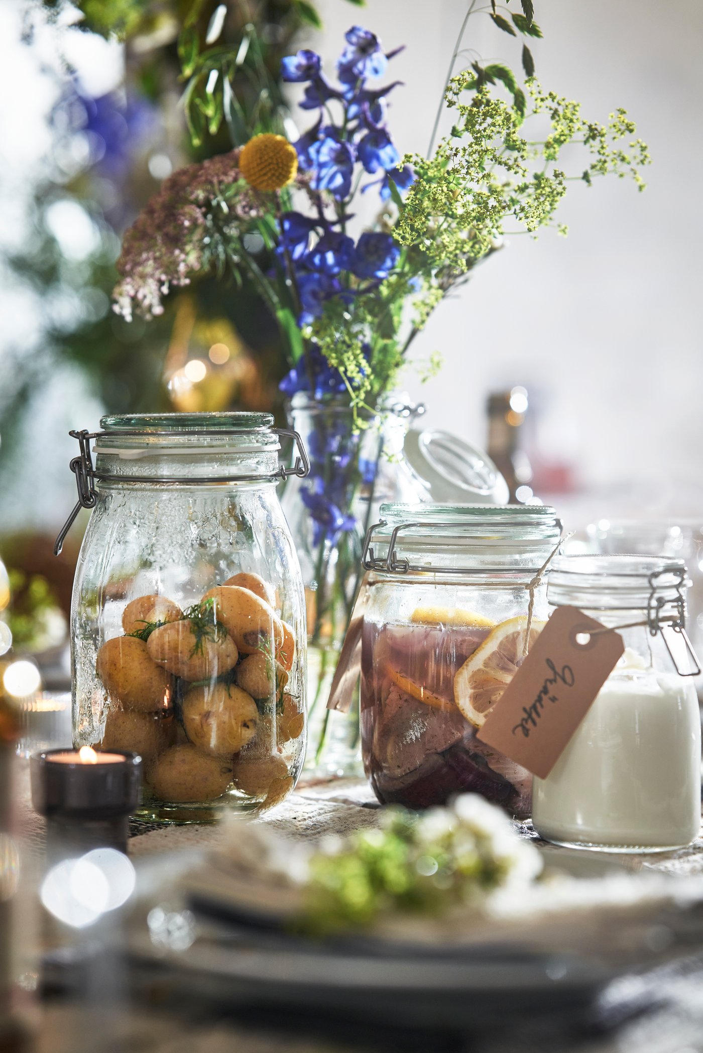 Three KORKEN glass jars with swing tops on a festively laid table. One contains potatoes, another a drink with oranges, and the third a white substance.