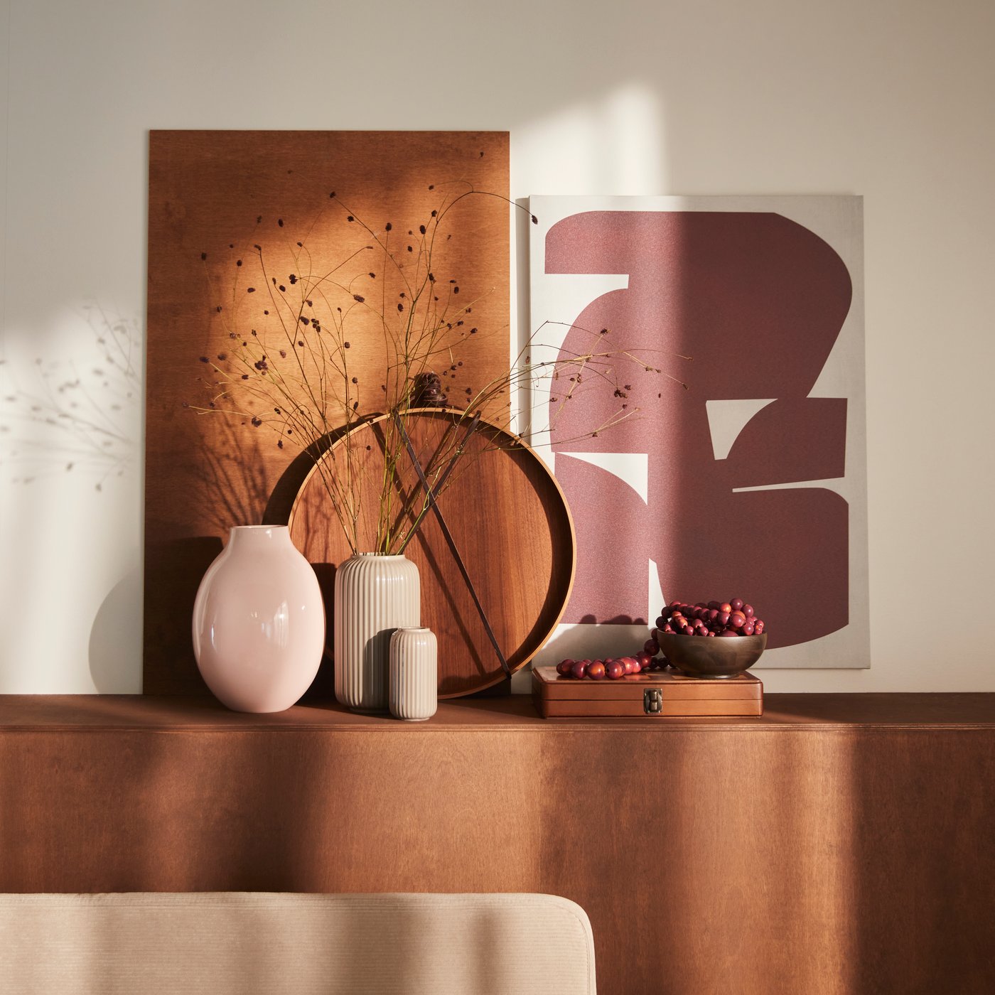 En sart pink BLODBJÖRK vase står på en gyldenbrun overflade sammen med flere vaser, dekorative genstande og et PJÄTTERYD billede.